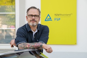 Ein Mann mit Brille und tätowiertem Unterarm lehnt an einem Auto und blickt in die Kamera; im Hintergrund ein gelbes Schild mit der Aufschrift "TÜV Rheinland FSP".
