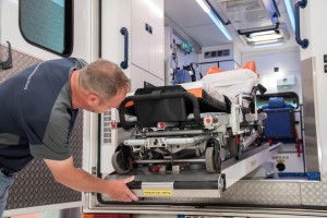 Ein Mann schiebt eine leere Trage auf einer ausziehbaren Rampe in einen Krankenwagen. Die Trage ist für Patiententransporte vorgesehen und das Innere des Rettungswagens ist sichtbar.