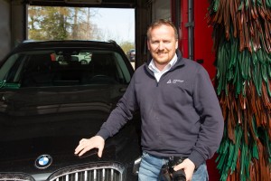 Ein lächelnder Mann steht in einer Waschanlage neben einem schwarzen BMW und hält eine Kamera in der rechten Hand; im Hintergrund sind rot-grüne Bürsten sichtbar.