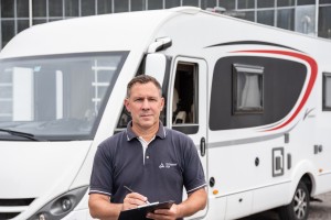 Mann im dunkelblauen Polohemd steht mit Klemmbrett und Stift vor einem weißen Wohnmobil, das im Hintergrund geparkt ist.