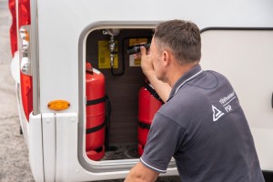 Ein Mann in grauem TÜV-Rheinland-Shirt prüft mit einer Taschenlampe zwei rote Gasflaschen in einem Seitenfach eines weißen Wohnmobils.