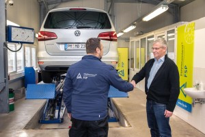 Ein Prüfer in TÜV Rheinland FSP-Jacke schüttelt einem Kunden die Hand in einer Werkstatt, im Hintergrund ein silberner VW auf einer Hebebühne und TÜV Rheinland FSP-Logos auf gelben Bannern.