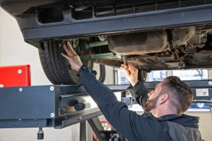 Ein Mann inspiziert das Fahrwerk eines Autos, das auf einer Hebebühne steht, und prüft mit einem Werkzeug die Bauteile unter dem Fahrzeug.