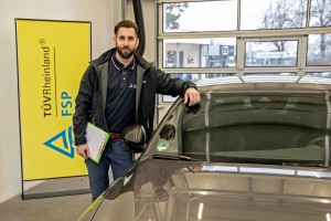 Ein Mann mit Bart steht in einer Werkstatt neben einem Auto, hält einen Klemmbrettblock in der linken Hand und lehnt sich mit der rechten Hand ans Autodach. Im Hintergrund steht ein gelbes Roll-up mit der Aufschrift „TÜV Rheinland FSP“.