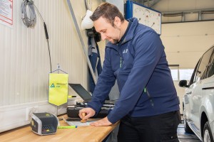 Ein Mann mit blauer Jacke stempelt ein Dokument auf einem Holztisch in einer Werkstatt. Im Hintergrund steht ein weißes Auto, an der Wand hängt ein gelbes TÜV Rheinland FSP-Schild. Auf dem Tisch liegen ein Kartengerät, ein Stift und weitere Unterlagen.
