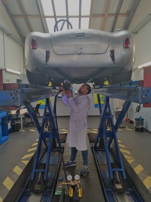 Ein Mechaniker im weißen Kittel arbeitet unter einem silbernen Oldtimer, der auf einer blauen Hebebühne in einer Werkstatt steht. Im Vordergrund stehen Werkzeuge und Messgeräte auf dem Boden.