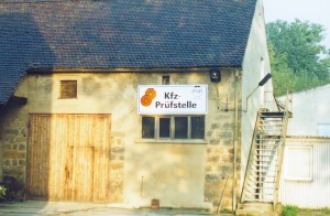 Altes Gebäude mit Holztor, Fenstern und Außentreppe. An der Fassade hängt ein Schild mit der Aufschrift „Kfz-Prüfstelle“ sowie zwei orangefarbenen Plaketten.
