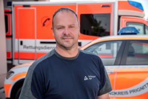 Ein Mann mit kurzem Haar und Bart steht vor einem Rettungswagen und einem Einsatzfahrzeug mit orangefarbener Markierung. Er trägt ein dunkelblaues T-Shirt mit TÜV Rheinland-Logo.