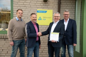 Vier Männer stehen lächelnd vor einem Gebäude mit gelbem TÜV Rheinland FSP Schild; zwei von ihnen geben sich die Hand, einer hält eine Urkunde.