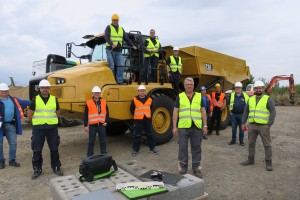 Gruppe von Männern in Warnwesten und Schutzhelmen posiert vor einem großen gelben Baustellenkipper im Freien; einige stehen, andere sitzen auf dem Fahrzeug, im Hintergrund sind weitere Baumaschinen zu sehen.