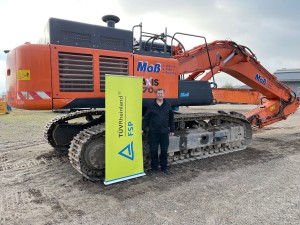 Ein Mann steht vor einem großen orangefarbenen Kettenbagger auf einem Schotterplatz. Neben ihm steht ein gelbes Roll-Up mit der Aufschrift „TÜV Rheinland FSP“. Der Himmel ist bewölkt.