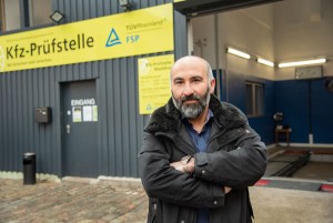 Ein Mann mit Bart steht mit verschränkten Armen vor einer Kfz-Prüfstelle mit gelbem TÜV Rheinland-Schild.