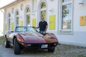 Ein Mann mit tätowierten Armen steht neben einem braunen Oldtimer-Cabrio vor einem weißen Gebäude mit großen Fenstern und gelben Schildern.