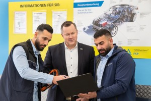 Drei Männer in Arbeitskleidung und Anzug stehen vor einer gelben Informationswand und betrachten gemeinsam einen aufgeklappten Laptop, im Hintergrund Schaubild zu Hauptuntersuchung eines Autos.