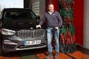 Ein Mann steht lächelnd neben einem schwarzen BMW-SUV in einer Waschanlage, hält eine Kamera in der Hand; hinter ihm ist eine bunte rot-grün-schwarze Waschbürste zu sehen.