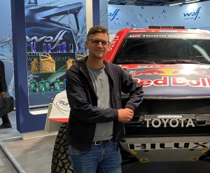 Ein Mann mit Brille steht lächelnd vor einem Rallyeauto mit Red Bull- und Toyota-Aufschrift, im Hintergrund ein Messestand mit Motoröl-Produkten von Wolf Lubricants.