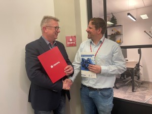 Zwei Männer stehen in einem Büroflur und geben sich die Hand, beide lächeln. Der Mann links hält eine rote Mappe mit dem Schriftzug "Equippo", der Mann rechts hält eine Broschüre mit einem blauen LKW auf dem Cover. Im Hintergrund ist durch Glas eine Büroeinrichtung mit Schreibtisch, Stuhl und Modell-Baumaschinen zu sehen.