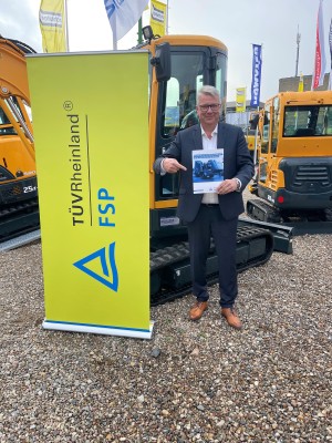 Ein Mann im Anzug steht auf einem Schotterplatz neben Baggern, hält ein Informationsblatt in der Hand und zeigt darauf. Links neben ihm steht ein gelbes Roll-Up mit dem Logo und Schriftzug von TÜV Rheinland FSP. Im Hintergrund befinden sich weitere Baumaschinen und Messefahnen.