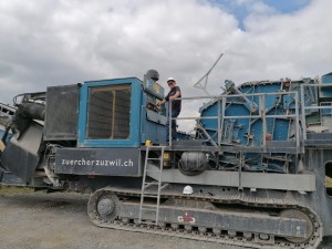 Ein Mann mit weißem Schutzhelm steht auf einer großen, blauen Baumaschine mit Kettenantrieb, im Hintergrund bewölkter Himmel; auf der Maschine steht die Webadresse "zuercherzuzwil.ch".