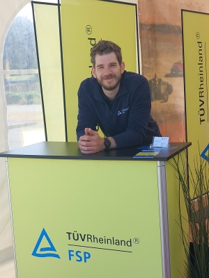 Ein Mann mit Bart und dunkler Kleidung lehnt lächelnd an einem gelben Informationsstand mit der Aufschrift „TÜV Rheinland FSP“. Im Hintergrund sind gelbe Aufsteller mit demselben Logo zu sehen.
