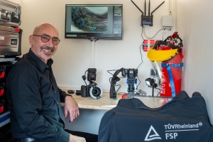 Ein lächelnder Mann mit Brille sitzt an einem Schreibtisch in einem Laborraum, umgeben von Kameras, technischem Equipment und einem Monitor, der ein vergrößertes Bild eines angebrannten Kabels zeigt. Im Hintergrund sind ein Feuerlöscher mit Gesichtsschutz und eine Jacke mit dem Aufdruck "TÜV Rheinland FSP" zu sehen.