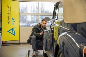 Ein Mann begutachtet aufmerksam einen Oldtimer in einer Halle; im Hintergrund steht ein gelbes Banner mit der Aufschrift "TÜV Rheinland FSP".