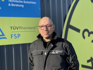 Ein Mann mit Brille und schwarzer Jacke mit der Aufschrift "TÜV Rheinland FSP" steht vor einem Gebäude mit Schildern von TÜV Rheinland FSP und einem großen gelben Prüfplaketten-Symbol.
