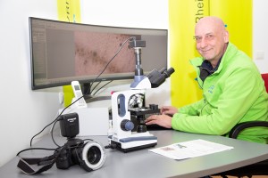 Ein Mann mit Glatze und grüner Jacke sitzt an einem Schreibtisch mit Mikroskop, Kamera und Monitor, auf dem eine vergrößerte Mikroskopaufnahme zu sehen ist.