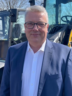 Mann mit grauen Haaren und Brille trägt ein weißes Hemd und einen dunkelblauen Anzug, steht im Freien vor einer Baumaschine; im Hintergrund sind Bäume ohne Blätter zu sehen.