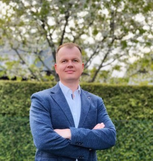 Mann mit kurzem Haar im blauen Sakko steht mit verschränkten Armen vor einer grünen Hecke und Bäumen; der Hintergrund ist unscharf.