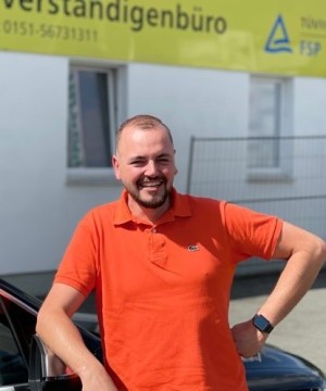 Ein Mann mit kurzem Bart und kurzem Haar trägt ein orangefarbenes Poloshirt und steht lächelnd mit einer Hand an der Hüfte neben einem schwarzen Auto. Im Hintergrund ist ein gelbes Schild mit der Aufschrift „Verständigengbüro“ und weiteren Textfragmenten sichtbar.