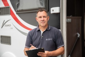 Ein Mann in dunkelblauem Polohemd mit TÜV-Rheinland-Logo steht mit Klemmbrett und Stift vor einem weiß-schwarzen Caravan mit rotem Streifen und der Aufschrift „Viseo i 700“.