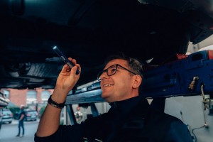 Ein Mann mit Brille inspiziert mit einer Taschenlampe die Unterseite eines Autos, das auf einer Hebebühne steht.