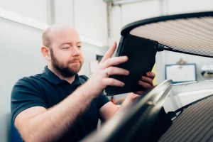 Ein Mann mit Bart und dunklem Polohemd inspiziert mit einem Tablet das geöffnete Heck eines Autos in einer Werkstatt.